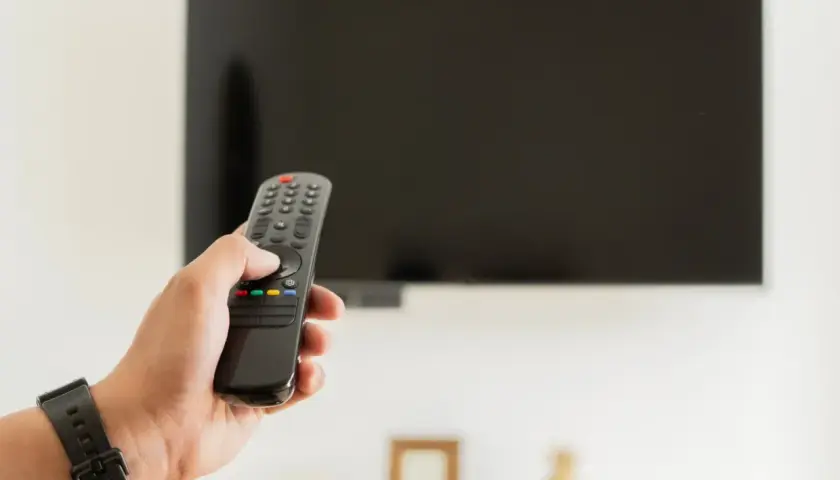 Dłoń trzymająca pilot do telewizora na tle wyłączonego ekranu.