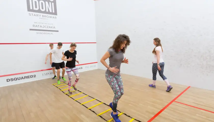 Grupa osób wykonuje dynamiczny stretching na drabince gimnastycznej w sali do squasha.