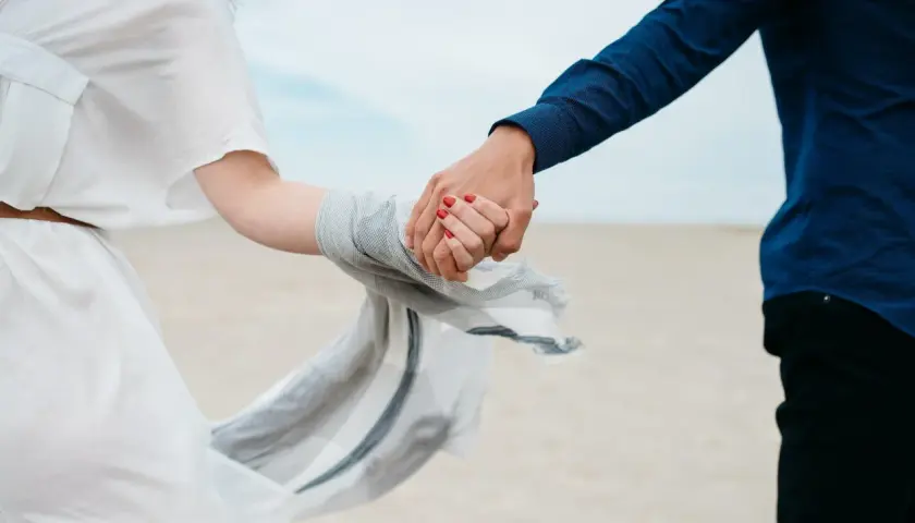 Dłonie splecione na plaży, symbol trwałych związków. Kobieta w zwiewnej białej sukience, mężczyzna w granatowej koszuli.