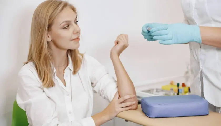 Młoda kobieta z blond włosami czeka na pobranie krwi. Pielęgniarka w niebieskich rękawiczkach trzyma probówkę. Badanie na czczo.