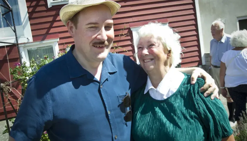 Sigge Dorsin ler mot kameran med en äldre kvinna. De står framför ett rött hus.