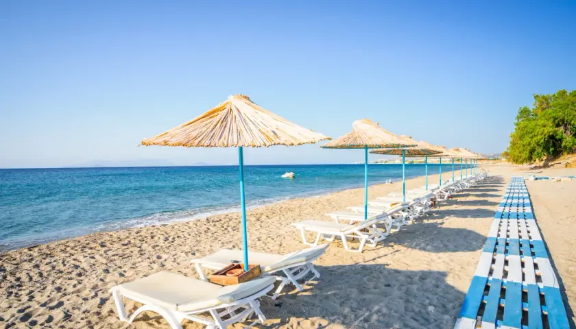 Słoneczna plaża na wyspie Kos, rzędy leżaków pod słomianymi parasolami, turkusowe morze i błękitne niebo. Idealne miejsce na relaks.