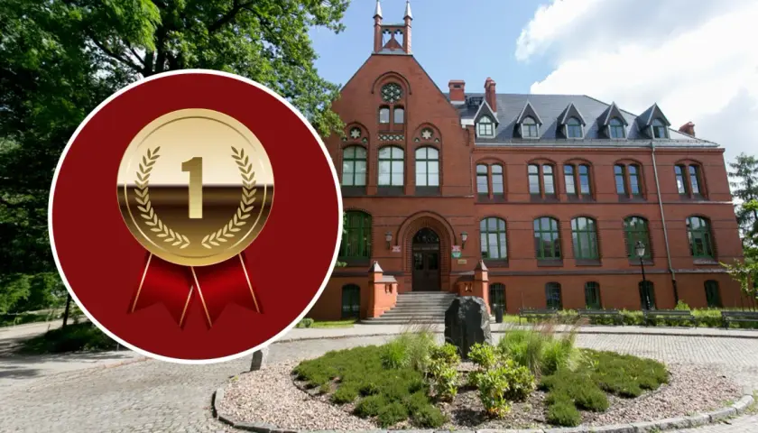 Złoty medal z cyfrą "1" na tle ceglanego budynku i zieleni.