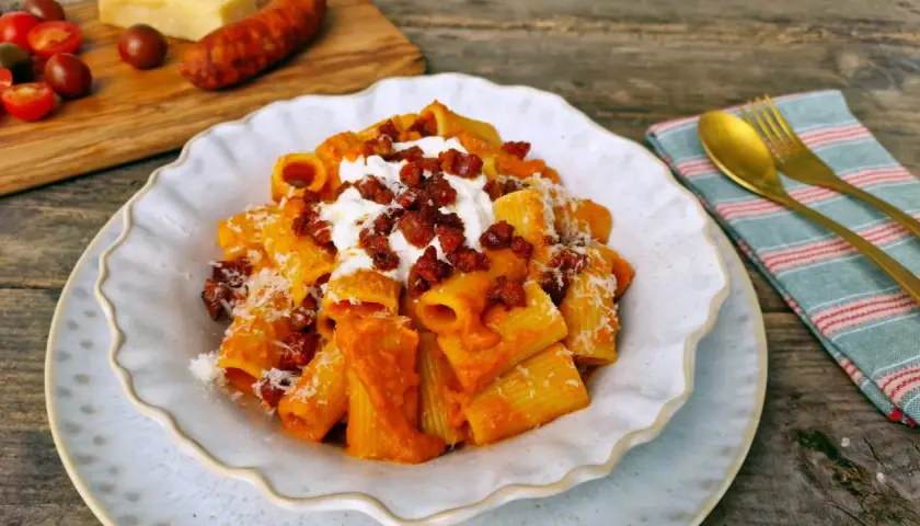 Un plato de macarrones con chorizo, cubiertos con queso rallado y una cucharada de crema. Al fondo, tomates y un trozo de queso.