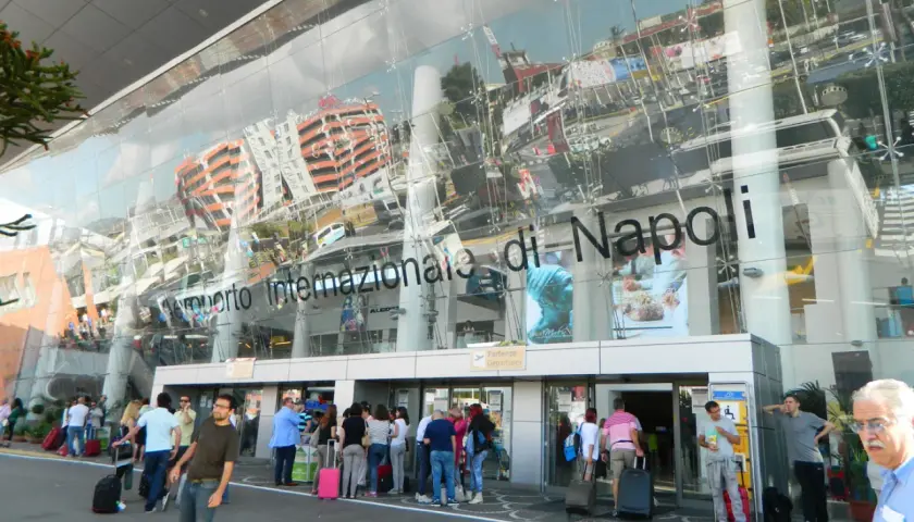 Ludzie z bagażami przed nowoczesnym budynkiem Aeroporto Internazionale di Napoli.