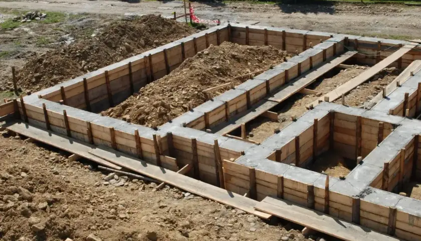 Budowa domu w toku: betonowe ławy fundamentowe w drewnianym szalunku, otoczone ziemią i gruzem.