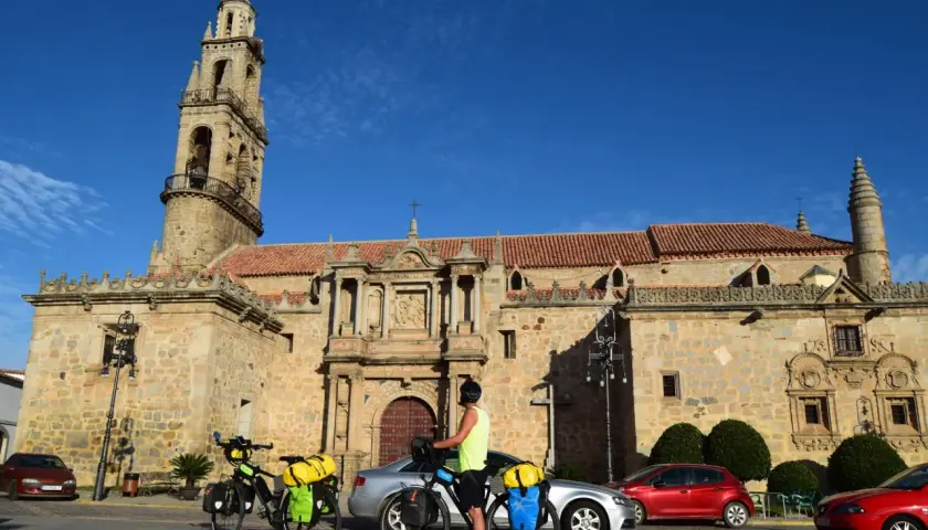 Ciclistas preparan sus bicicletas para continuar las etapas de Transandalus frente a una iglesia histórica bajo un cielo azul.