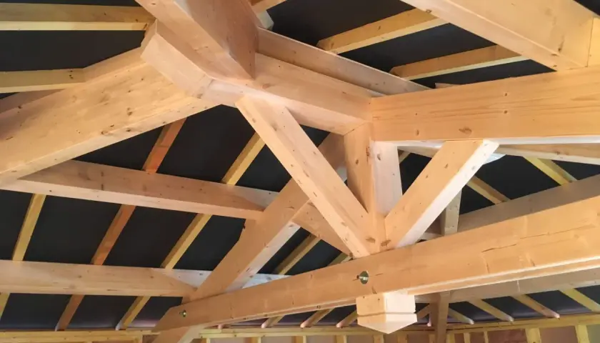 Vue rapprochée d'une charpente traditionnelle 2 pans en bois clair, avec des poutres massives et des chevrons assemblés.