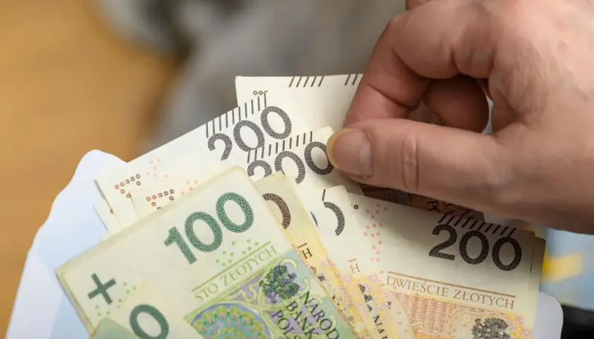 Dłoń trzyma plik banknotów 100 i 200 zł, symbolizujący nową płacę minimalną.