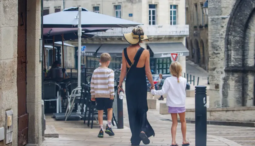Kobieta w letniej sukience i kapeluszu prowadzi dwójkę dzieci, chłopca i dziewczynkę, ulicą. To francuskie dziecko, które zwiedza miasto.
