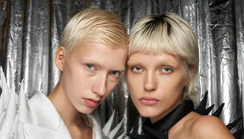 Zwei Models mit kurzen, blonden Haaren und blassen Gesichtern vor silbrigem Hintergrund.