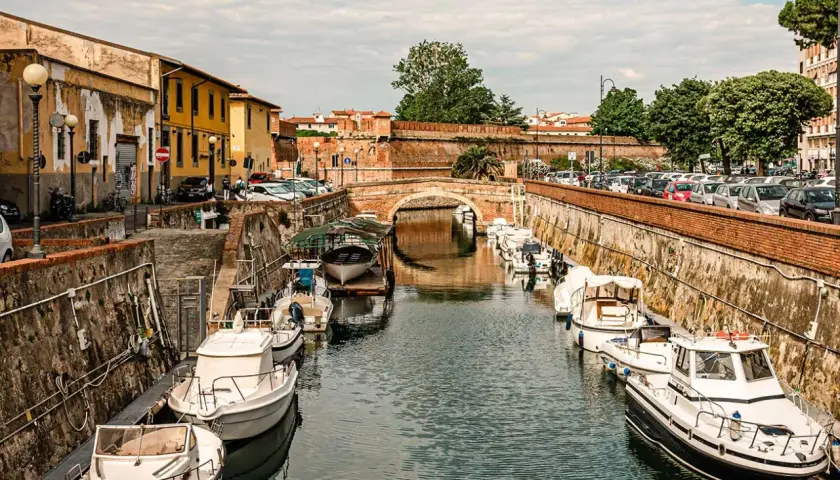 Boats docked along a canal in Livorno, Italy, with a brick bridge in the background.