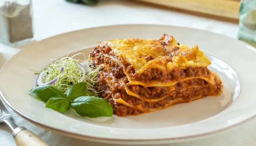 Porción de lasaña de carne casera, con capas de pasta, salsa boloñesa y queso gratinado, servida con brotes y albahaca.