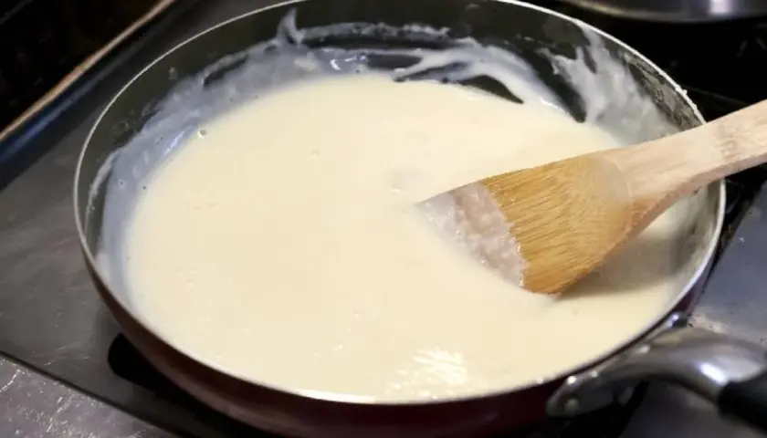 Une spatule en bois remue une sauce onctueuse dans une poêle. La préparation crémeuse, une sauce vin blanc et crème, est prête à être servie.