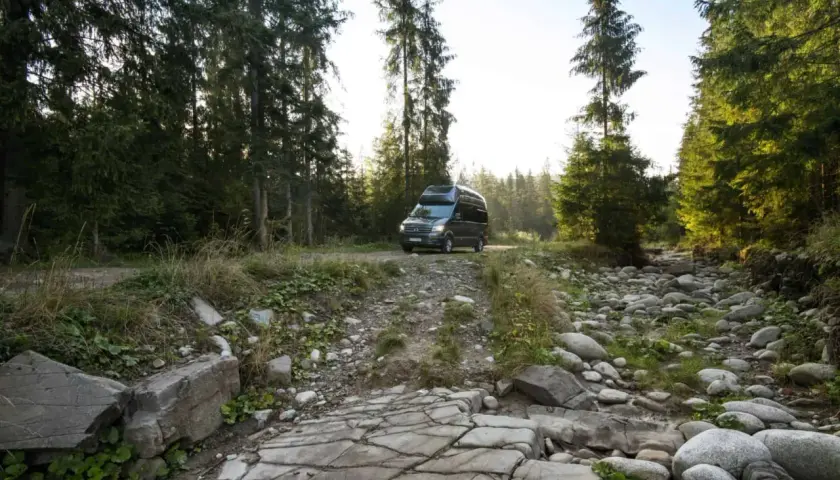 Ciemny kamper na leśnej drodze, otoczony przez wysokie drzewa i kamieniste koryto strumienia. Idealne miejsce na **split camping** i ucieczkę od cywilizacji.