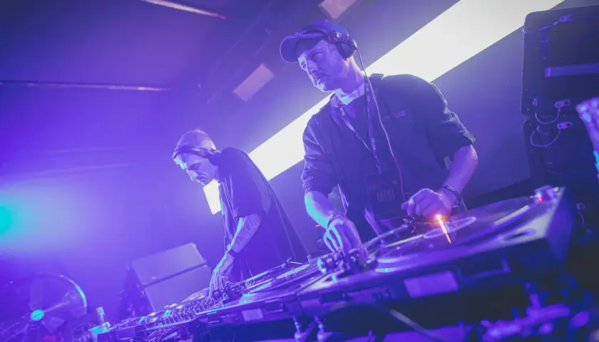 Dwóch DJ-ów w słuchawkach tworzy muzykę techno. Jeden z nich ma na szyi identyfikator "ARTIST".