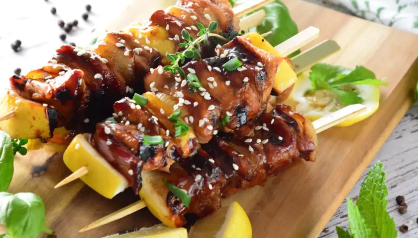 Aromatyczne yakitori z kurczaka w sosie teriyaki, posypane sezamem i szczypiorkiem, podane z cytryną na drewnianej desce.