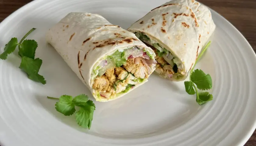 Kebab w tortilli przekrojony na pół, z kurczakiem, warzywami i ziołami, na białym talerzu.
