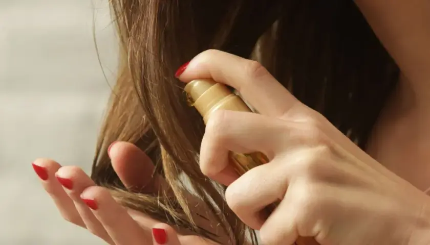 Eine Hand mit roten Nägeln sprüht ein goldenes Öl auf trockene Haare, um sie zu pflegen.