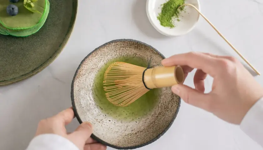 Hände bereiten Matcha zu, ein gesunder Genuss für die matcha schwangerschaft. Daneben grüne Pancakes mit Blaubeeren.