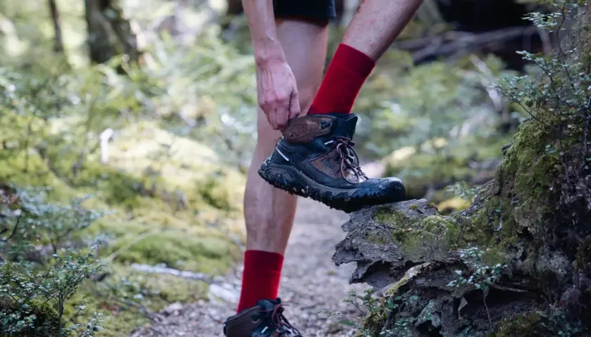 A hiker adjusts their muddy Keen shoes, showing signs of wear and tear, as they step onto a moss-covered log in a forest.
