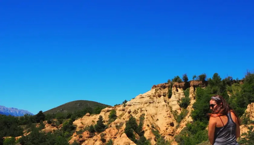 Una mujer contempla el paisaje de la Cerdanya, con sus formaciones rocosas ocres y vegetación verde bajo un cielo azul intenso.