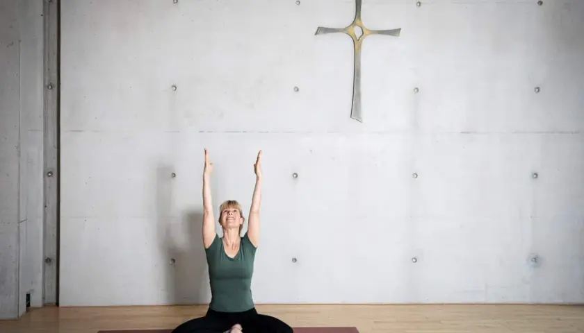Frau in Yoga-Pose mit erhobenen Armen vor einem Kreuz an einer Betonwand. Eine harmonische Verbindung von Yoga und Christentum.
