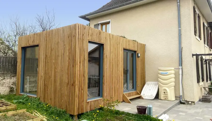 Une extension en bois moderne s'intègre à une maison existante, avec de grandes fenêtres et une finition en lattes de bois.