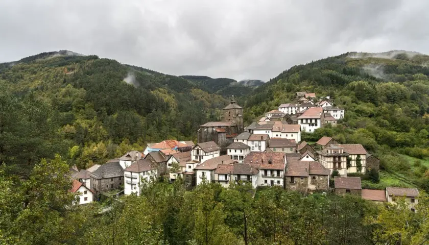 Pueblo pintoresco en el Pirineo Navarro, con casas de piedra y tejados rojos, rodeado de bosques frondosos y montañas nubladas.