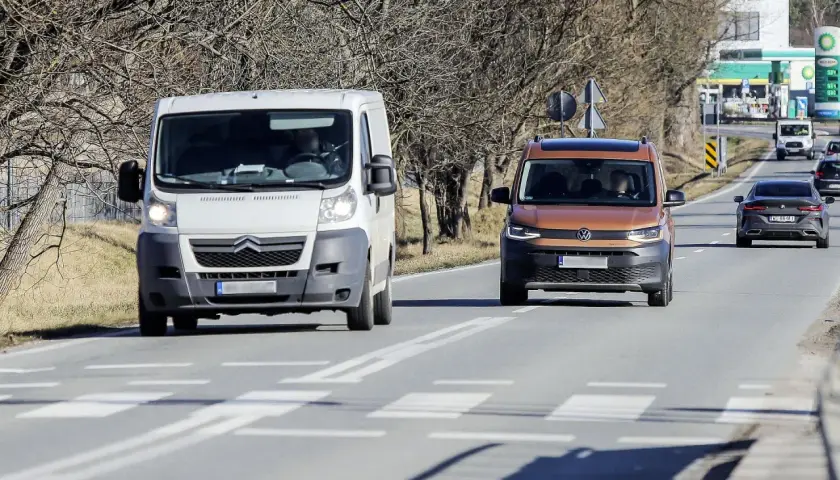 Biała furgonetka wyprzedza na przejściu dla pieszych pomarańczowego vana. W tle stacja benzynowa.