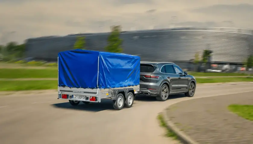 Szary SUV ciągnie dwuosiową przyczepę pod niebieską plandeką. Czy można ciągnąć przyczepę dwuosiową na kat. B?