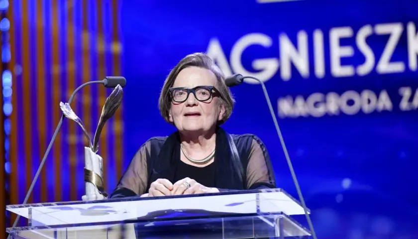 Agnieszka Holland w okularach za mównicą z mikrofonem i statuetką.
