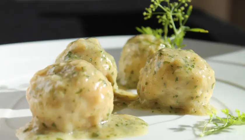 Quattro polpette in umido, ricoperte da una salsa cremosa alle erbe, servite su un piatto bianco con guarnizione di prezzemolo.