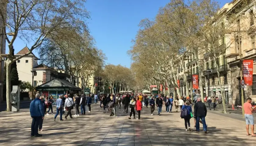 Tłum ludzi spaceruje po słonecznej La Rambla w Barcelonie, otoczonej drzewami i zabytkowymi budynkami.