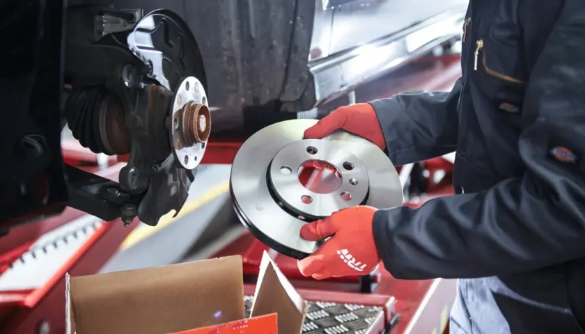 Mechanik w czerwonych rękawiczkach trzyma nową tarczę hamulcową obok zamontowanej tarczy.