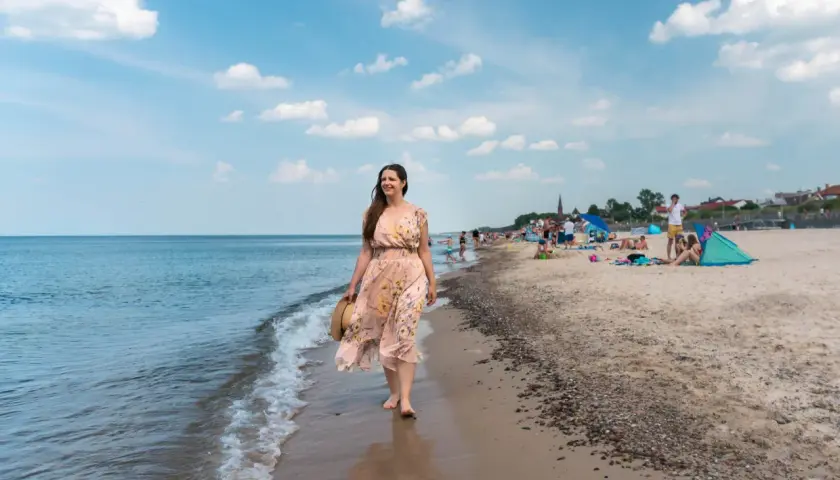 Kobieta w kwiecistej sukience spaceruje brzegiem morza. W tle plaża pełna ludzi, parawanów i domków – typowe sarbinowo atrakcje.