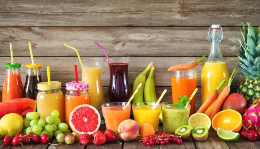 Varietà di bevande colorate e freschi estratti di frutta, con frutta intera e a fette su uno sfondo di legno.