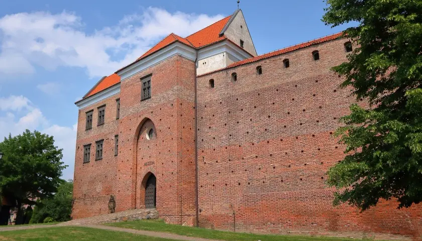 Zamek w Łęczycy, ceglana twierdza z czerwonej cegły, z charakterystycznym łukowym wejściem i oknami, otoczona zielenią.