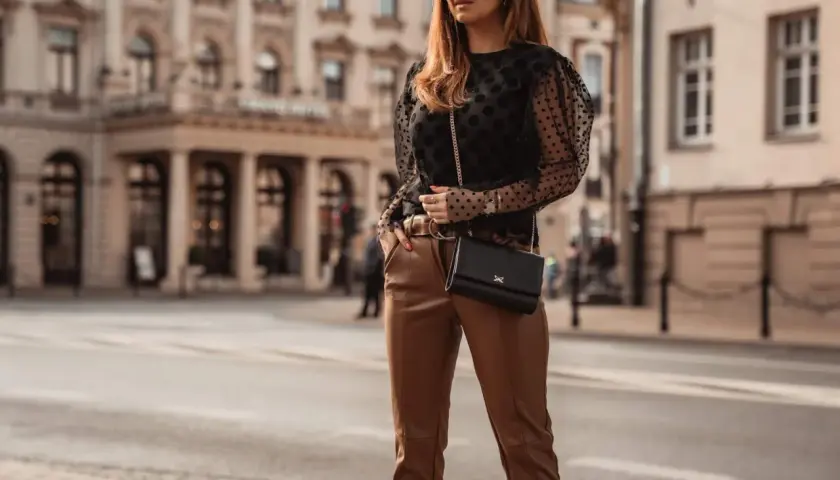 Elegancka kobieta w brązowych spodniach i czarnej bluzce w kropki, z czarną torebką na łańcuszku, pozuje na tle miejskiej kamienicy.