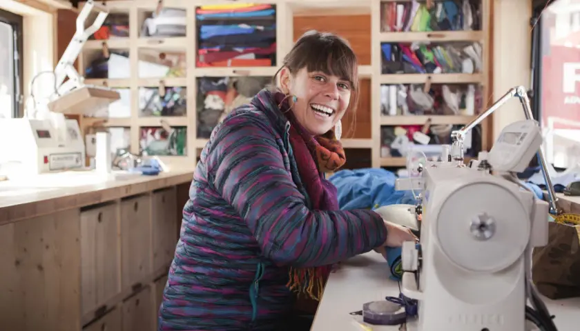 Frau lacht fröhlich an ihrer Nähmaschine, bereit für die Patagonia Reparatur. Regale voller bunter Outdoor-Kleidung im Hintergrund.