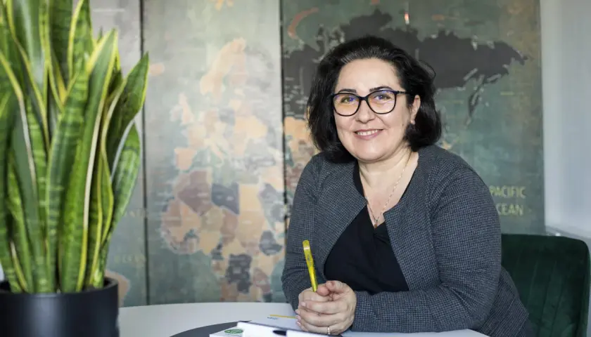 Frau mit Brille lächelt, hält Stift. Im Hintergrund Weltkarte. Sie berät zu Lohnsteuerhilfeverein Kosten.