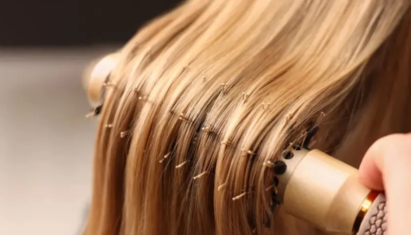 Delikatne rozczesywanie blond pasm przedłużanych włosów okrągłą szczotką. Poznaj sekrety, jak dbać o przedłużane włosy, by były lśniące i zdrowe.