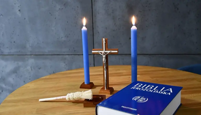 Dwie niebieskie świece, krzyż, kropidło i Biblia Jerozolimska na drewnianym stole. Symbolika religijna, jakby przygotowanie do kolędy.