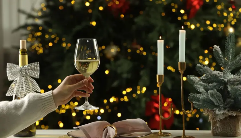 Une main tient un verre de vin blanc pétillant, parfait pour un buffet froid. Une bouteille décorée et des bougies illuminent la scène devant un sapin de Noël scintillant.