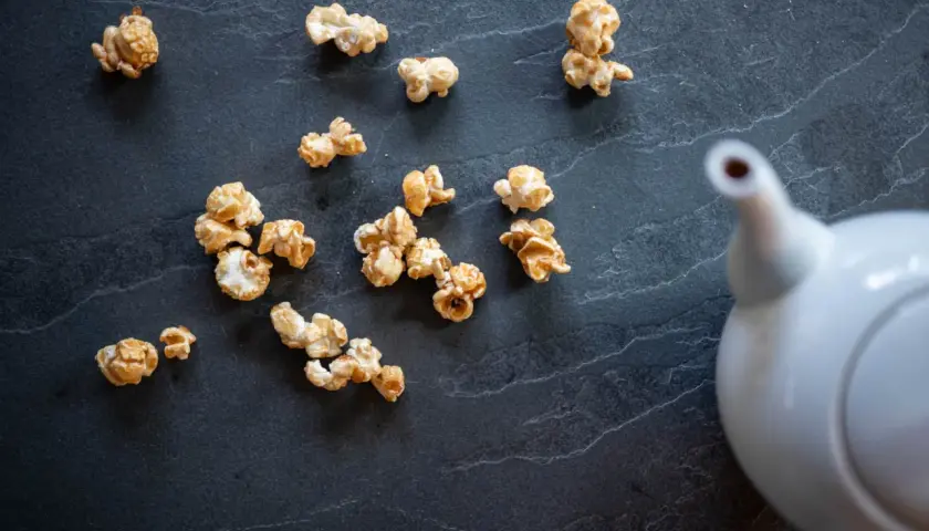 Pop corn caramel éparpillé sur une surface sombre, avec une théière blanche en arrière-plan.