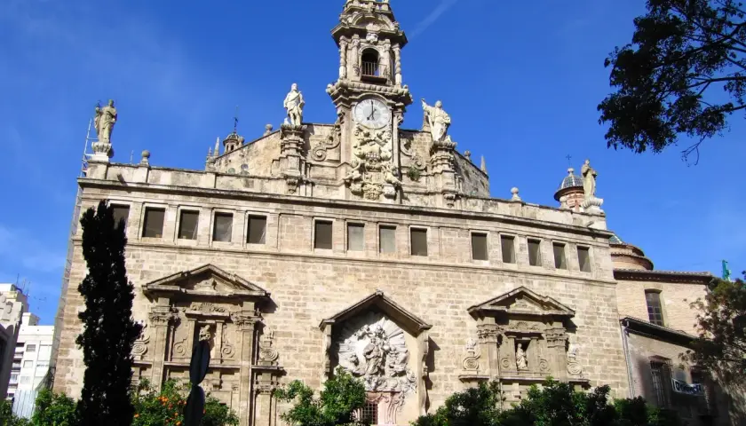 La **iglesia de los Santos Juanes**, con su imponente fachada de piedra, torre con reloj y estatuas, se alza majestuosa bajo un cielo azul intenso.