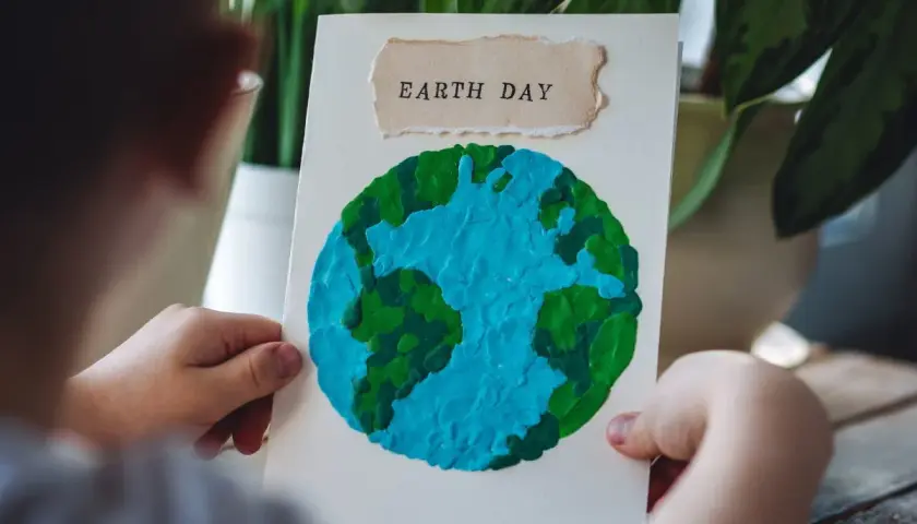 Dziecko trzyma kartkę z pracą plastyczną przedstawiającą planetę Ziemię z plasteliny. Napis "EARTH DAY" na górze.