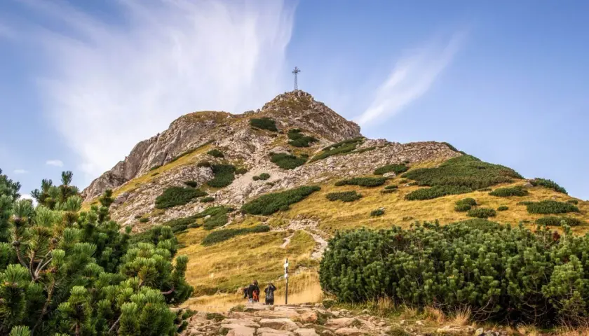 Kamienista ścieżka prowadzi pod górę, gdzie widać krzyż na szczycie. Turyści idą w stronę, by zobaczyć, ile ma Giewont.