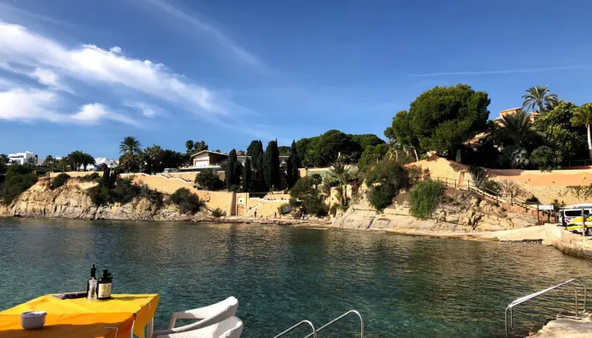 Mesa con mantel naranja y sillas blancas junto al mar cristalino en cala les Bassetes. Casas y vegetación en la ladera.