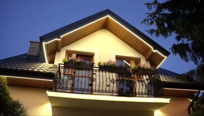 Elegancki dom z oświetleniem led w podbitce, podkreślającym architekturę. Balkon z kwiatami i ozdobną balustradą.